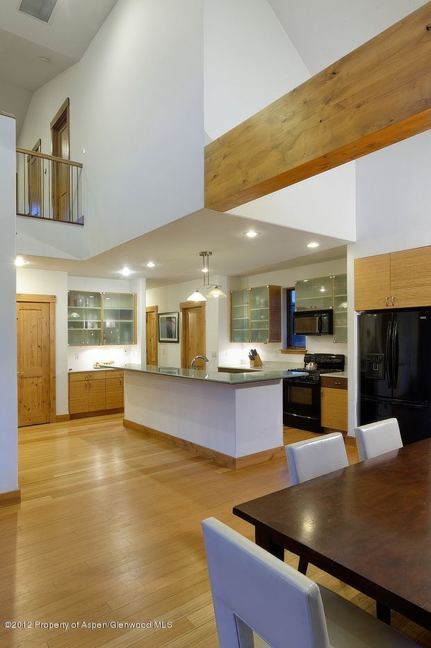 A bright open-plan kitchen and dining area with a large central island, pale wood flooring, light wood cabinetry, glass-fronted upper cabinets, stainless fixtures, and black appliances. The space has a dramatic double-height ceiling with exposed timber beams and an upper-level balcony or landing with timber railings.