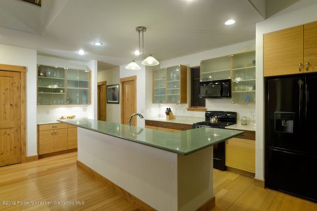A spacious, warm-toned kitchen with a large central island featuring a pale green glass benchtop and white waterfall-style sides. The room includes extensive light timber cabinetry, glass-front upper cabinets, stainless-steel appliances, a black refrigerator, wood flooring, recessed ceiling lights, and pendant lights over the island.