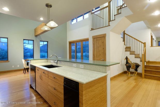 Open-plan kitchen and circulation space with a large central island, pale stone benchtops, timber cabinetry, integrated sink, pendant lighting, and adjoining dining area. The room features light timber flooring, pale walls, tall windows, exposed timber accents, and a staircase with dark metal balustrades leading to an upper level.