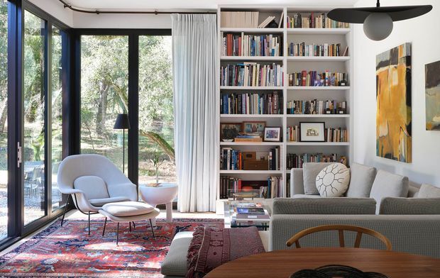 A stylish, light-filled living room with floor-to-ceiling black-framed glazing opening toward a leafy outdoor setting. The room features extensive built-in white shelving filled with books, a grey sofa and armchairs, a sculptural black pendant light, colourful abstract artwork, a large patterned rug, and a timber dining table in the foreground. The space combines contemporary architecture with curated mid-century-inspired furnishings.
