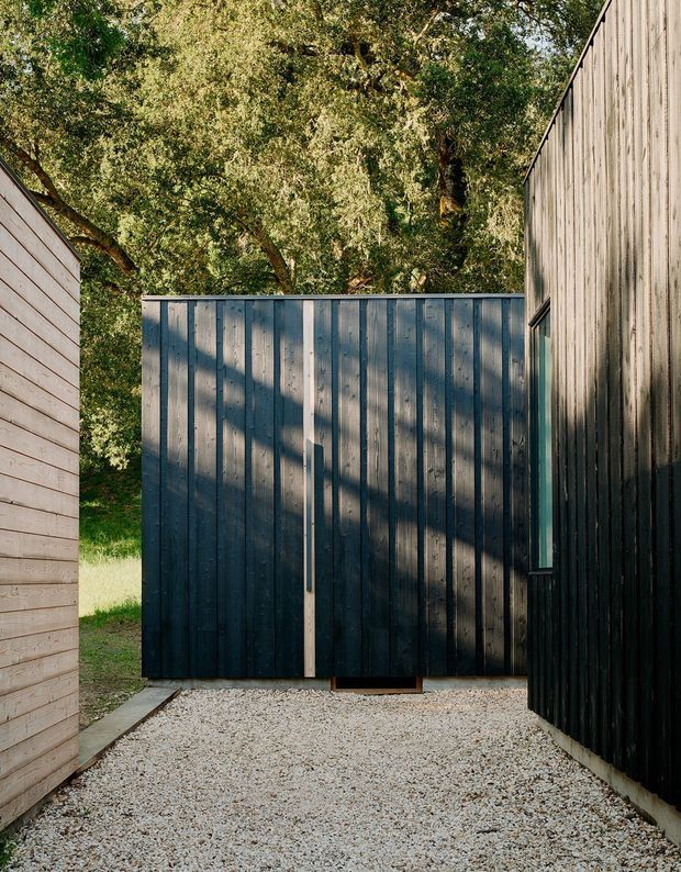 A narrow gravel passage between compact architectural structures, with dark vertically clad timber walls and a matching enclosed gate or facade ahead. A lighter horizontal timber-clad wall frames the left side, while mature trees and dappled sunlight are visible beyond. The composition emphasizes contrasting timber cladding, simple geometric forms, and a sheltered outdoor circulation space.