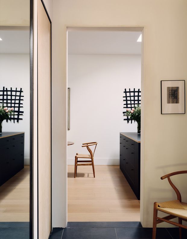 A view through a doorway into a refined minimalist interior, showing a light timber-floored room with dark built-in cabinetry on both sides, pale walls, a woven-seat timber chair, abstract wall art, and a black grid-like wall feature with floral arrangement. The foreground has dark tiled flooring and a second timber chair partially visible. The composition emphasizes the transition between spaces and a clean, restrained material palette.
