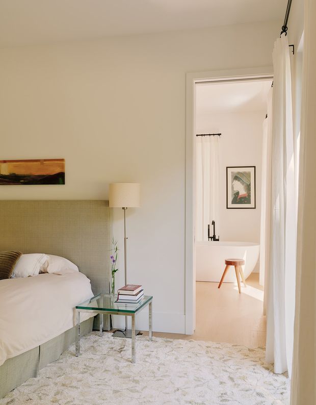 Bright, softly styled bedroom with a pale upholstered bed and textured green headboard, white bedding, glass bedside table, slim floor lamp, and a large light patterned rug. Tall white curtains frame a doorway into a minimalist bathroom, where a freestanding white bathtub, stool, wall art, and black tapware are visible. The room has warm neutral walls and abundant natural light.
