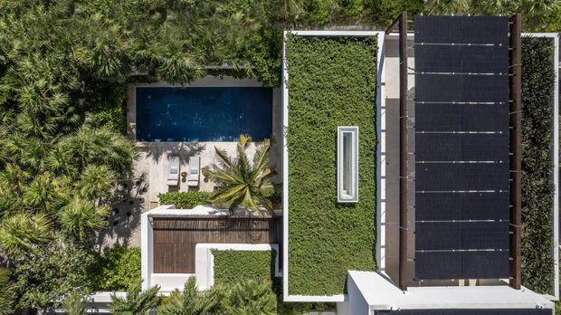 Aerial view of a modern residential property surrounded by dense tropical landscaping. The composition shows a rectangular swimming pool with a pale paved deck, outdoor loungers, a narrow landscaped green roof or planted roof section, and a large dark linear roof or pergola structure with a skylight-like opening. The architecture is largely concealed by vegetation, with strong geometric forms and a lush resort-like setting.