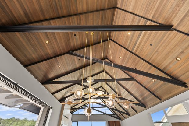 Upward-looking interior view of a dramatic pitched timber ceiling with exposed dark structural beams and black metal bracing. Recessed spotlights and a cluster of suspended globe pendant lights hang beneath the high ceiling. Large glazed openings at the sides and end bring in daylight and provide views of the outdoors.
