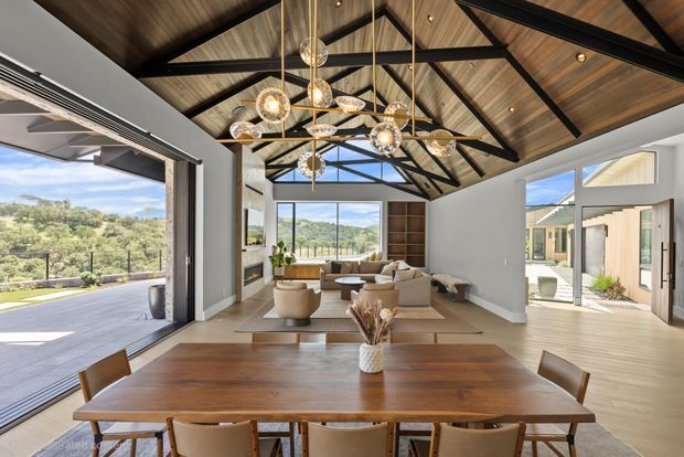 A bright, spacious open-plan dining and living area with a large timber dining table in the foreground, opening to a contemporary lounge. The room features extensive floor-to-ceiling glazing and wide-open doors to an outdoor terrace, a dramatic vaulted timber ceiling with exposed dark structural beams, and a statement cluster of spherical pendant lights. Built-in timber shelving, pale flooring, neutral furnishings, and expansive landscape views create an airy indoor-outdoor setting.