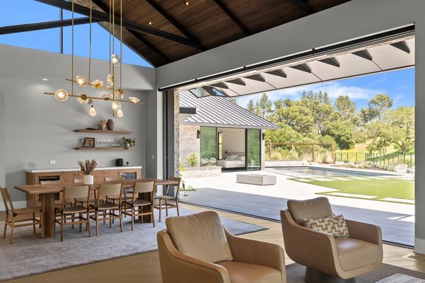 A sophisticated open-plan dining and living area with a large retractable glazed opening connecting directly to a landscaped outdoor terrace and pool area. The interior features a timber-lined pitched ceiling, dark structural framing, warm timber flooring, a wooden dining table with upholstered chairs, tan lounge chairs, minimalist shelving, and a sculptural suspended pendant light. Outside, a paved terrace, lawn, pool, and a small pavilion are visible.