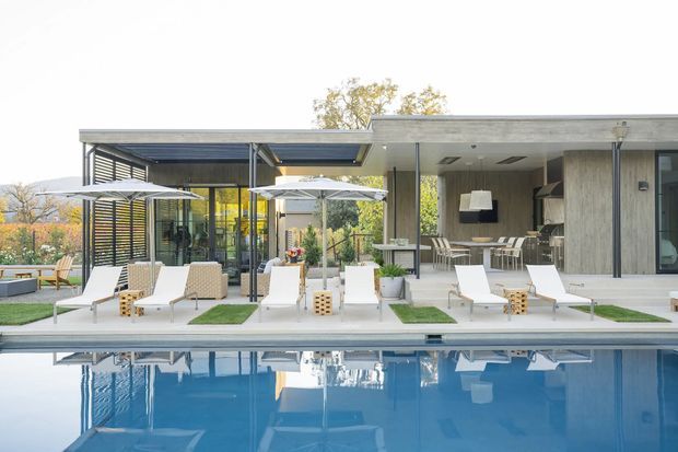 A contemporary single-storey residence viewed from the pool, featuring a broad flat-roofed pavilion, extensive floor-to-ceiling glazing, slender dark structural posts, and a covered outdoor entertaining area. White loungers and umbrellas line the pool terrace, while a concrete-toned outdoor kitchen or bar with seating is visible beneath the roof.