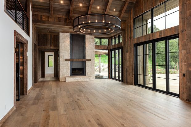 A spacious, high-ceilinged living room with exposed timber beams and a timber-planked ceiling, wide-plank wood flooring, and large black-framed floor-to-ceiling glazing. A substantial pale stone fireplace forms the focal point, while a large circular multi-light pendant hangs centrally. The room has a warm, rustic architectural character with contemporary glazing and detailing, and opens visually to a green outdoor setting.
