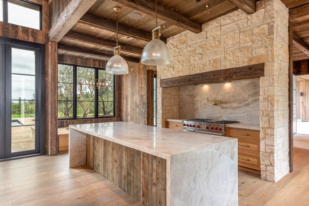 Rustic-modern kitchen with a large pale stone island, timber-clad island base, exposed heavy timber beams and ceiling boards, natural stone masonry walls, dark-framed floor-to-ceiling windows, and stainless-steel pendant lights. A built-in range and timber cabinetry sit within a stone feature wall, with a glimpse of an adjoining room beyond.