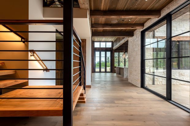 Interior view of a contemporary open-plan circulation and living space featuring a floating timber staircase with black metal balustrade, exposed timber ceiling beams, wide timber flooring, pale stone masonry walls, and large black-framed glazed doors and windows. The corridor opens toward bright exterior views, creating a warm, spacious, industrial-modern atmosphere.