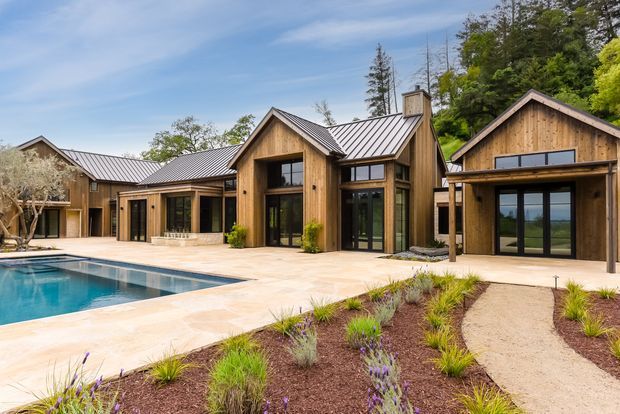 A sprawling single-storey residential compound arranged around a swimming pool and paved terrace. The buildings feature warm vertical timber cladding, steep gabled forms, dark standing-seam metal roofs, large black-framed glazed doors and windows, and prominent timber beams and entry canopies. Native-style landscaped planting and gravel paths occupy the foreground, while the pool and outdoor entertaining area dominate the courtyard-like setting.