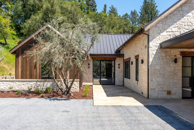 Exterior view of a low-rise, multi-wing building set into a wooded, sloping site. The facade combines pale rough-cut stone masonry with dark timber cladding, a standing-seam metal gable roof, black-framed glazing, and a covered entry courtyard. Gravel paving, light stone paving, landscaped planting, and mature trees surround the entrance.