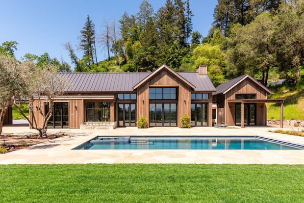 Exterior view of a single-storey timber-clad house set against a wooded hillside. The building has multiple gabled roof volumes, dark metal roofing, large black-framed glazed doors and windows, a paved pool terrace, and a rectangular swimming pool in the foreground.