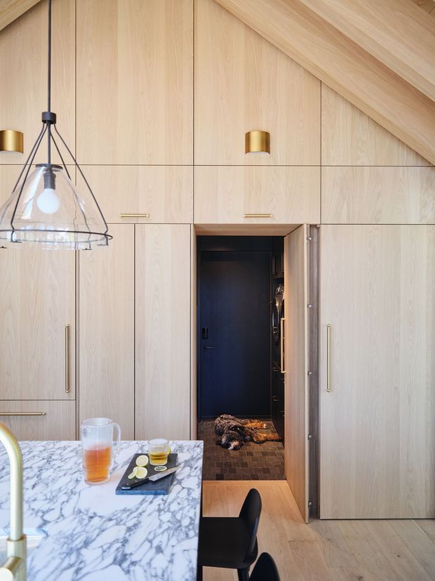 A warm, light-filled kitchen with extensive pale timber floor-to-ceiling cabinetry and integrated storage. A veined white marble island occupies the foreground, with a sculptural glass pendant light above it. Brass-toned handles and wall lighting add subtle contrast. An open concealed door reveals a dark tiled pantry or utility space, while the sloping timber-lined ceiling and matching timber floor create a cohesive, refined interior.