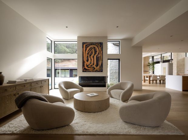 A refined open-plan living room with a sculptural arrangement of curved upholstered lounge chairs around a round timber coffee table on a large cream rug. A linear fireplace with a prominent framed artwork forms the focal point, while a long timber sideboard lines the left wall. Floor-to-ceiling glazing brings in natural light and views of greenery, and the adjacent dining and kitchen areas are visible through the open-plan layout. The scheme is warm, neutral, contemporary, and understated.