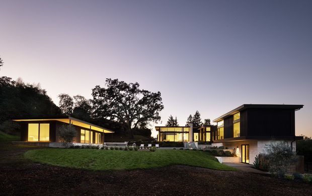 A dusk exterior view of a low-slung modern residential property arranged across a gently sloping landscaped site. Several flat-roofed volumes with dark cladding, extensive warm-lit floor-to-ceiling glazing, and connecting terraces surround a central outdoor area. Mature trees and lawn frame the composition.