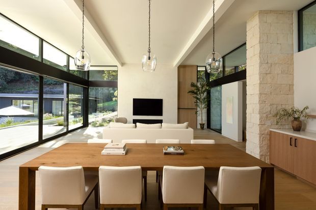 A bright open-plan living and dining area with a long solid timber dining table and upholstered dining chairs in the foreground, and a neutral sofa with wall-mounted television beyond. Floor-to-ceiling black-framed glazing wraps the room, bringing in garden views. A pale stone feature wall, timber cabinetry, white walls, and pendant glass lights create a refined contemporary interior.