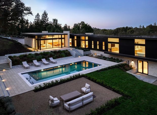 A wide dusk view of a large, low-slung modern residence arranged around a landscaped courtyard and rectangular swimming pool. The house features strong horizontal flat-roofed forms, dark exterior cladding, pale rendered or masonry walls, extensive warm-lit glazing, and broad paved terraces. Outdoor lounge furniture, poolside loungers, lawn, planting beds, and gravel areas create a refined indoor-outdoor entertaining setting.
