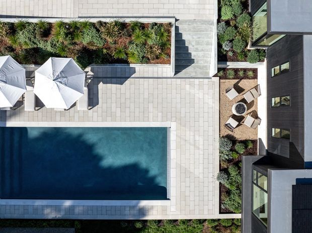 Top-down aerial view of a contemporary residential courtyard featuring a rectangular swimming pool, pale paved terraces, outdoor umbrellas and seating, landscaped garden beds, and dark modern building wings surrounding the space.