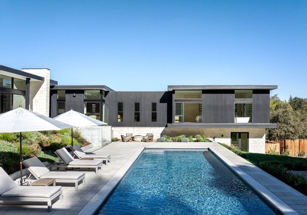 Contemporary multi-wing residence viewed from the landscaped pool terrace, with a long rectangular swimming pool, pale stone paving, lounge chairs and umbrellas in the foreground. The house has a flat-roofed composition, dark vertical cladding, extensive glazing, light masonry feature walls, and a clean geometric arrangement set against mature planting and a clear blue sky.