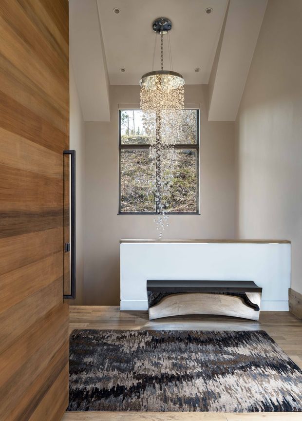 A refined residential entryway with a dramatic crystal pendant light suspended from a recessed ceiling. The space features a large horizontal-grain timber pivot door with black hardware, warm timber flooring, pale neutral walls, a white built-in console with a sculptural timber inset, and a large abstract grey, black, and cream rug. A tall black-framed window brings in natural light and overlooks vegetation.