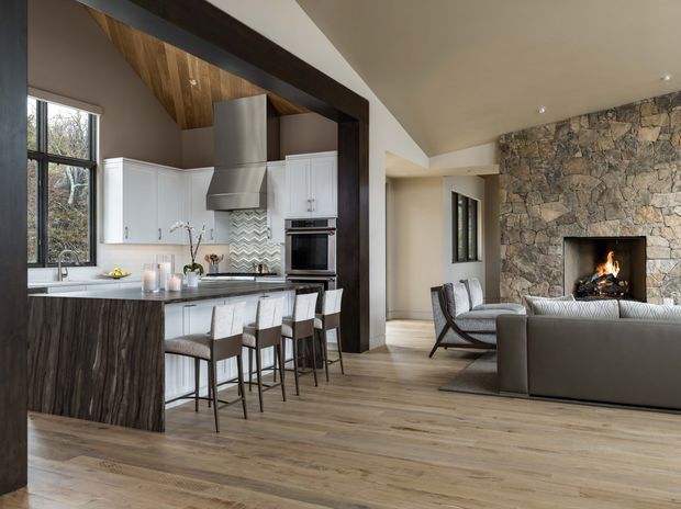 Open-plan contemporary kitchen and living room with a large dark timber island, white shaker-style cabinetry, stainless steel appliances, a patterned tile splashback, and a warm wood-lined ceiling detail. The adjacent lounge features neutral upholstered seating and a substantial rough-stone fireplace wall with a lit fire. Light timber flooring, dark structural beams, and large windows create a warm, refined interior.