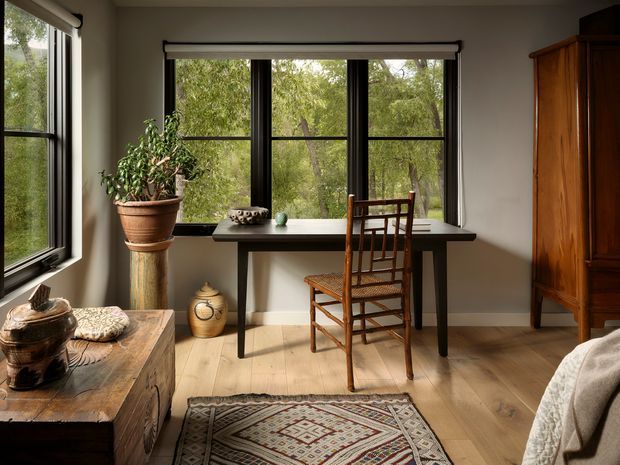 A warm, light-filled bedroom or sitting space with large black-framed windows overlooking trees. A narrow desk or console is positioned beneath the central window with a wooden ladder-back chair, while a substantial wooden armoire, rustic chest, potted plant, ceramic vessels, and patterned rug add earthy character. The room has pale timber flooring, soft neutral walls, and a calm natural outlook.