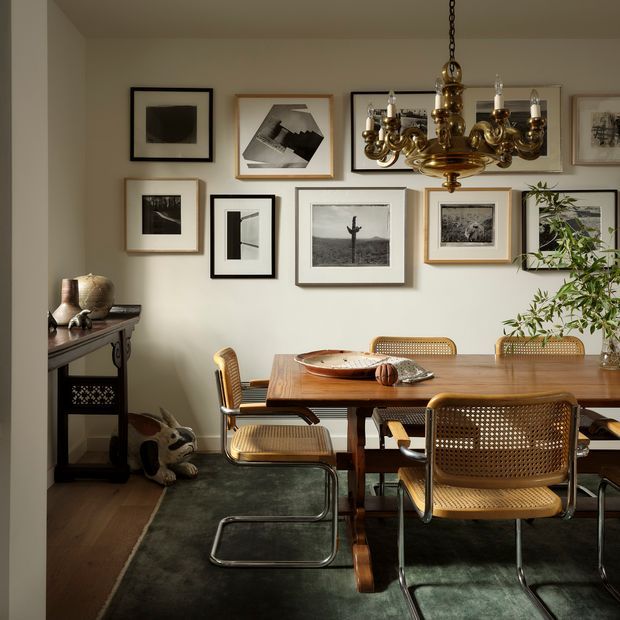 A warmly styled dining room with a large wooden dining table surrounded by woven cane and tubular metal chairs. A vintage brass chandelier hangs above the table, while a gallery wall of framed black-and-white artwork decorates the light neutral wall. A dark console table, sculptural decor, timber flooring, and a deep green area rug complete the refined interior.