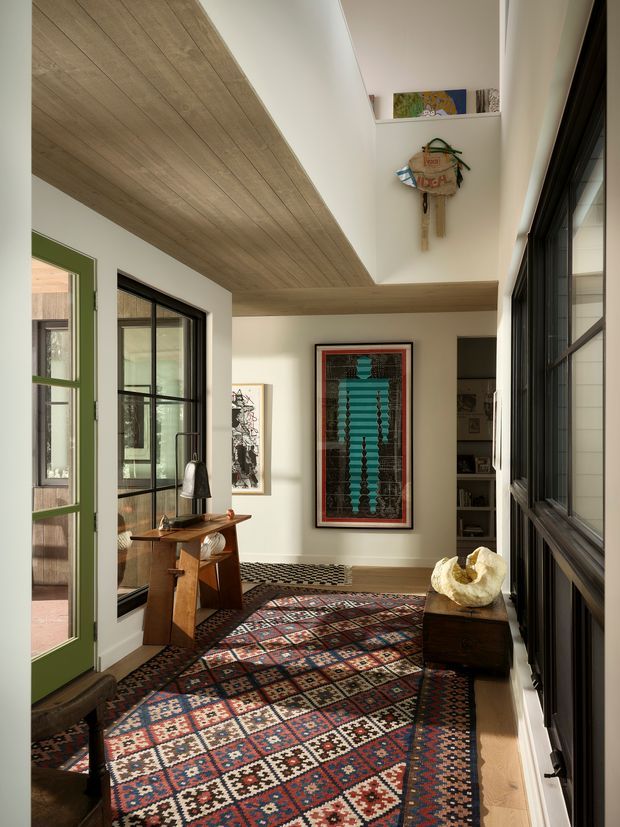 A bright, narrow interior passage or landing with extensive black-framed glazing on both sides, a green-framed glazed door, and a warm timber-lined ceiling forming a strong horizontal soffit. The space is furnished with a small timber console and stool, layered patterned rugs, framed artwork, and a dark timber chest with sculptural decor. A high recessed shelf displays artwork and woven objects. The overall design combines crisp white walls, natural timber, dark metal frames, and richly coloured textiles.