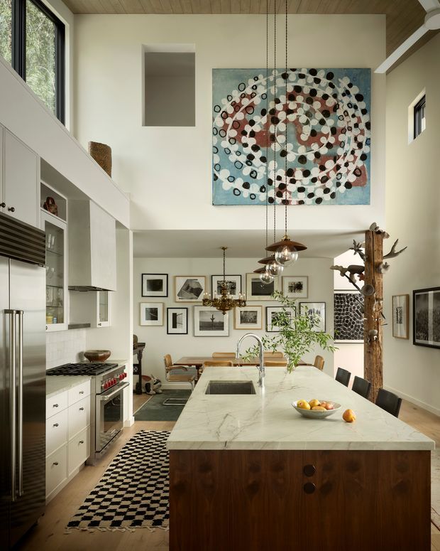 A spacious open-plan kitchen and dining area with a dramatic double-height ceiling. The room features white built-in cabinetry, stainless-steel appliances, a large veined stone island with dark timber cabinetry, pendant lights, timber ceiling boards, extensive wall art, and a sculptural natural timber post. A dining table and chairs are visible beyond the island, with abundant natural light from tall windows.
