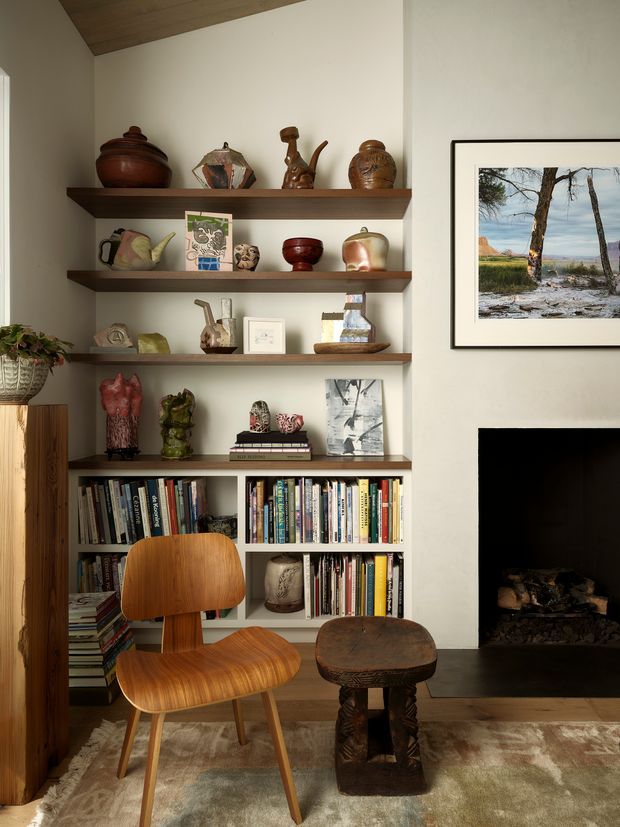 Warm, eclectic living room vignette with built-in open shelving filled with books, ceramics, sculptures and decorative objects. A mid-century-style wooden lounge chair and carved wooden coffee table sit on a large neutral rug beside a dark fireplace. The room has cream walls, rich timber shelving and ceiling boards, and framed artwork.