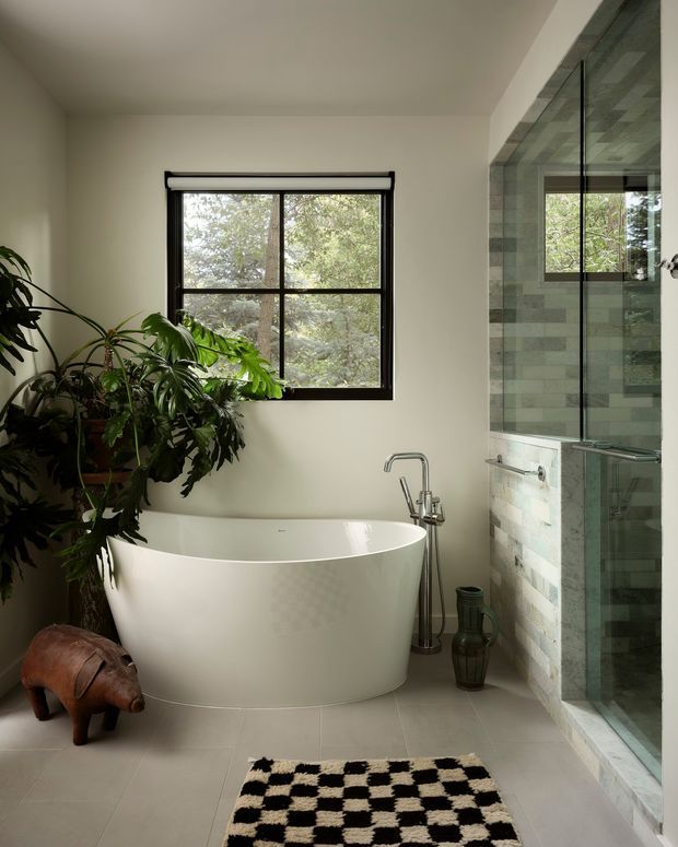 A bright, softly styled bathroom featuring a freestanding white oval bathtub beneath a black-framed window, a wall-mounted tub filler, and a large glass-enclosed shower clad in pale grey-green rectangular tiles. Lush indoor plants sit beside the tub, with a monochrome checkerboard rug and sculptural dark accessories adding contrast. The room has a calm, natural, contemporary feel.