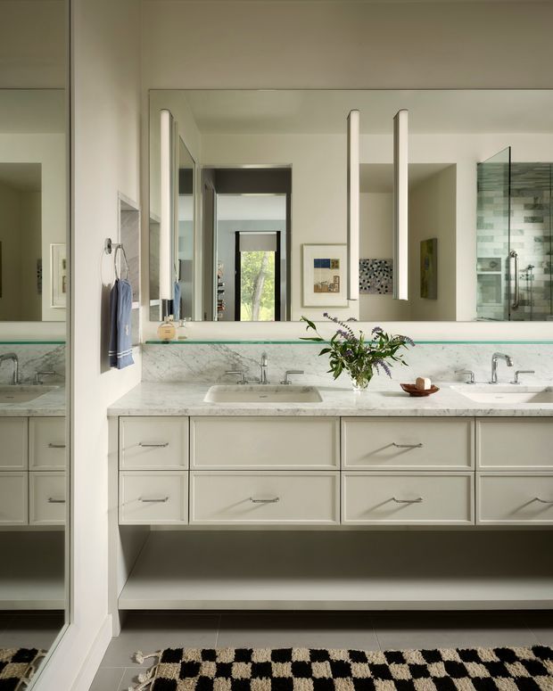 A bright, refined bathroom centered on a long double vanity with pale framed cabinetry, a light marble countertop, two integrated basins, and chrome tapware. A large mirrored wall reflects the adjoining space, while a glass-enclosed shower with pale stone or tiled walls is visible at right. The room features white walls, dark floor tiles, hanging towels, framed artwork, and a black-and-cream patterned rug.
