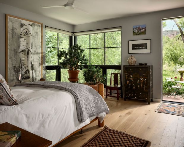 A warm, light-filled bedroom with a large bed in the foreground, pale timber flooring, grey walls, and expansive black-framed corner glazing overlooking greenery. The room includes a tall abstract artwork, indoor plants, a dark decorative cabinet, a wooden chair, layered rugs, and a glazed door opening to an outdoor sitting area. The overall character is eclectic and contemporary with natural materials and strong indoor-outdoor connection.