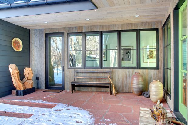 Covered outdoor entry or courtyard with dark horizontal cladding, weathered timber wall and ceiling lining, black-framed glazing and a glazed door. The space has a terracotta-tiled floor, built-in timber bench shelving, sculptural wooden chairs, large ceramic vessels and a bright green-framed opening at right. Snow is visible beyond the patio, while trees and the surrounding landscape appear through the glazing.