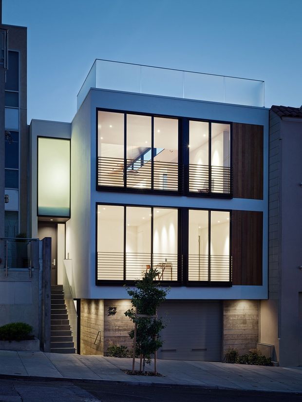 Twilight exterior view of a narrow multi-storey urban townhouse or apartment-like residence. The contemporary facade combines a white rendered box form, extensive black-framed floor-to-ceiling glazing, horizontal metal balcony railings, dark vertical timber-look panels, and a lower garage integrated into the street frontage. A narrow entry stair rises beside the building, with neighbouring attached buildings visible on both sides.