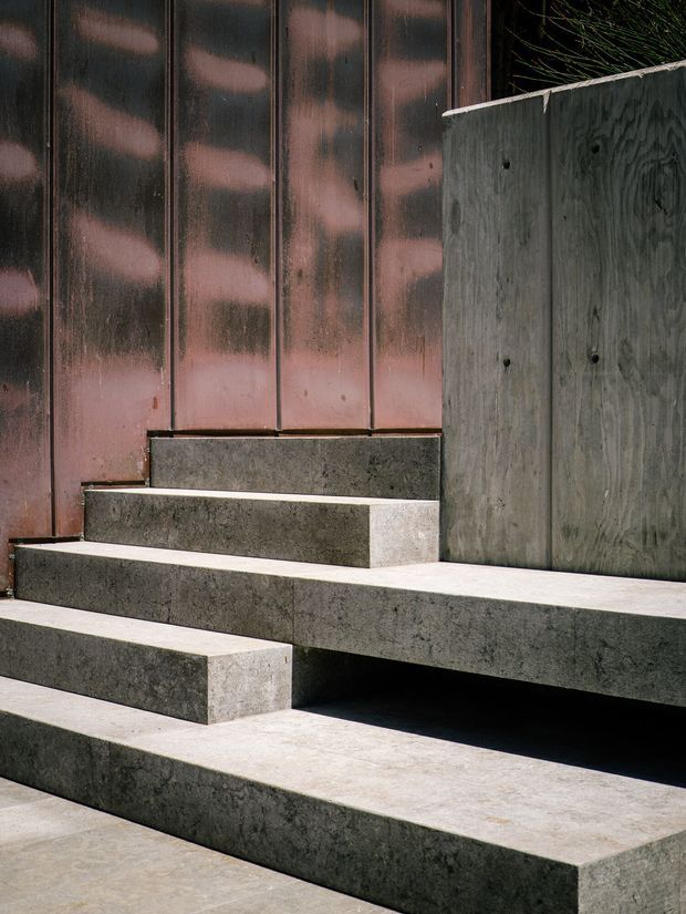 Close-up architectural view of broad stepped concrete seating or stairs beside a dark vertically ribbed metal-clad wall and a raw plywood panel. The composition emphasizes layered horizontal slabs, heavy materiality, and strong light-and-shadow contrast in a minimalist exterior or semi-exterior setting.
