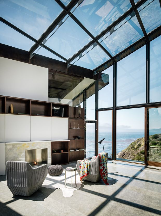 A bright, double-height living room with expansive floor-to-ceiling glazing and a glazed roof structure overlooking the ocean and rugged coastline. The space features dark timber built-in shelving and cabinetry, a pale stone fireplace surround, polished concrete flooring, sculptural upholstered lounge chairs, and strong geometric daylight shadows. The interior has a contemporary, refined coastal character with an indoor-outdoor connection.