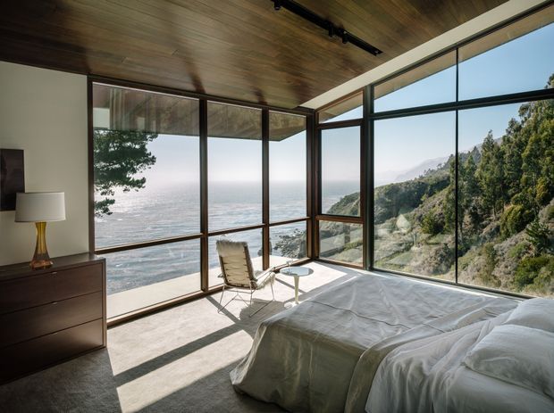 A serene bedroom with a low-profile bed, dark timber ceiling, and expansive floor-to-ceiling corner glazing overlooking the ocean and a steep, tree-covered coastal landscape. The room includes a dark wood dresser, table lamp, lounge chair, and small round side table, with strong natural light and an unobstructed panoramic outlook.