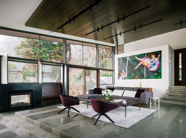 A spacious contemporary living room with floor-to-ceiling glazing, a dark stone fireplace, low modular sectional sofa, sculptural burgundy lounge chairs, and a large area rug. The room features a dramatic timber-lined ceiling with black track lighting, polished stone flooring, broad stair steps, and a colourful artwork on the wall. The extensive glazing overlooks mature vegetation and brings abundant natural light into the space.