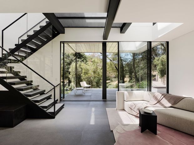 A bright, open-plan contemporary living room with a dramatic black steel staircase, exposed dark structural beams, polished concrete flooring, and expansive floor-to-ceiling black-framed glazing opening toward a leafy outdoor setting. A large neutral sectional sofa, pale rug, and minimalist side table occupy the foreground. The high ceiling and glazed rear wall create an airy, refined interior with strong indoor-outdoor connection.
