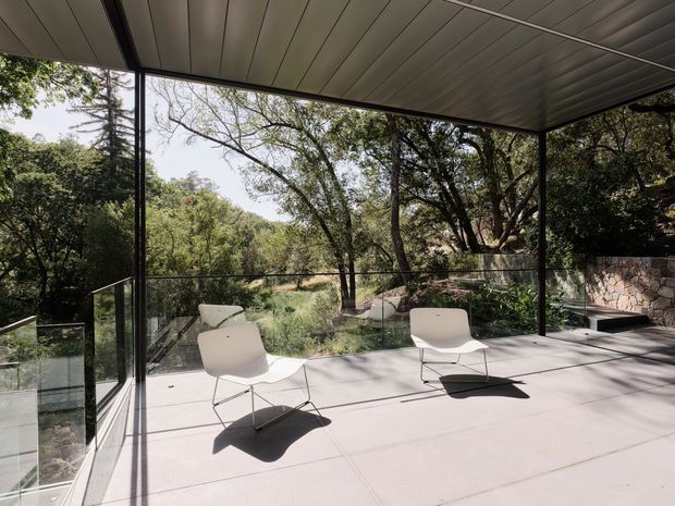 A spacious covered outdoor terrace or balcony with a sleek dark metal-framed structure, full-height glazing, transparent glass balustrades, and a large pale tiled or concrete floor. Two white modern lounge chairs sit in the foreground, overlooking a wooded natural landscape. A rough stone retaining wall and dense mature vegetation reinforce the indoor-outdoor connection. The composition is minimalist and contemporary, with strong linear shadows and abundant filtered daylight.