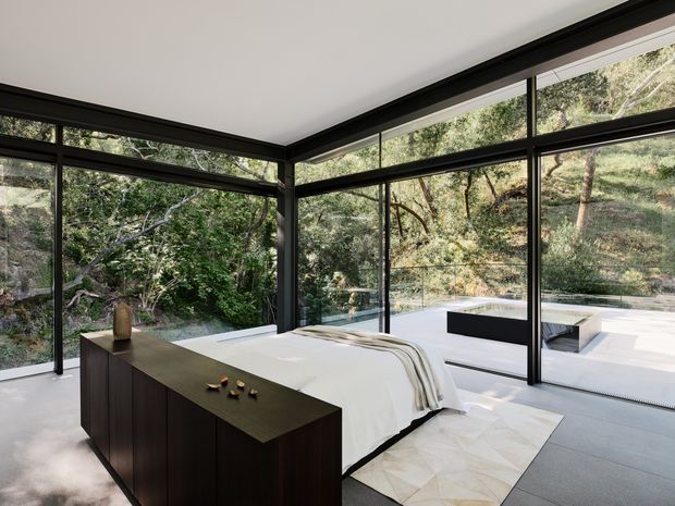 A contemporary bedroom with a low-profile bed positioned beside extensive floor-to-ceiling black-framed glazing. A dark wood storage cabinet sits at the foot of the bed, while the room opens visually and physically to a paved outdoor terrace surrounded by trees. The space has a restrained, minimalist composition with a neutral palette and strong connection to the landscape.