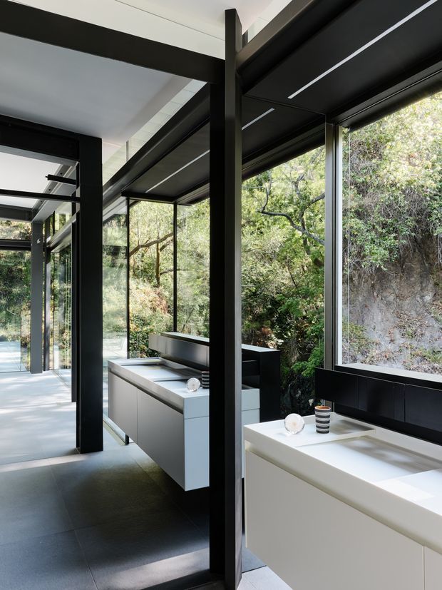 A sleek contemporary kitchen interior with a long white island or counter in the foreground, dark built-in cabinetry and a secondary grey kitchen island beyond. Floor-to-ceiling glazing runs along the room, opening views to dense greenery and a rocky bushland setting. Strong black structural posts and beams frame the glazing, with integrated linear ceiling lighting and dark large-format flooring creating a refined, high-contrast composition.
