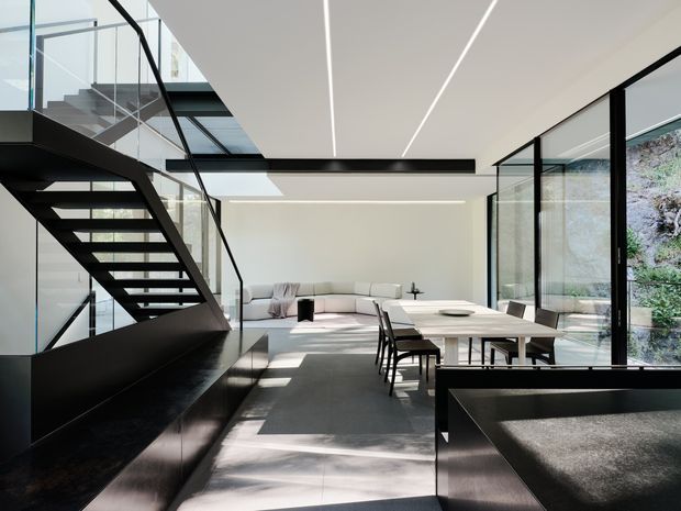 A bright, open-plan contemporary interior combining a dining area and living room beneath a dramatic black open-riser staircase. Full-height glazing, glass balustrades, linear recessed ceiling lights, pale flooring, and black built-in joinery create a crisp, minimalist composition. A long light dining table sits beside a pale sectional sofa, with greenery visible through the windows.