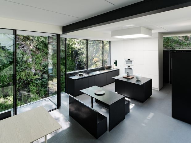 A contemporary open-plan kitchen with dark matte cabinetry, a central island, integrated appliances, and a compact dining table. Full-height black-framed glazing wraps the room and overlooks dense greenery, while a large rectangular pendant provides focused illumination. The palette is monochrome and restrained, with pale flooring and walls contrasting against black joinery.