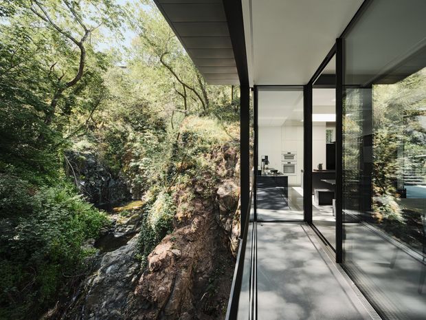 A contemporary glass-walled extension or corridor projects over a wooded rocky ravine with a small flowing stream. The image looks along a narrow covered walkway with dark slim-framed floor-to-ceiling glazing on the right, revealing a bright minimalist kitchen or living area inside. The structure uses strong horizontal and vertical lines, a flat roof or soffit, and a close connection to the surrounding bushland.