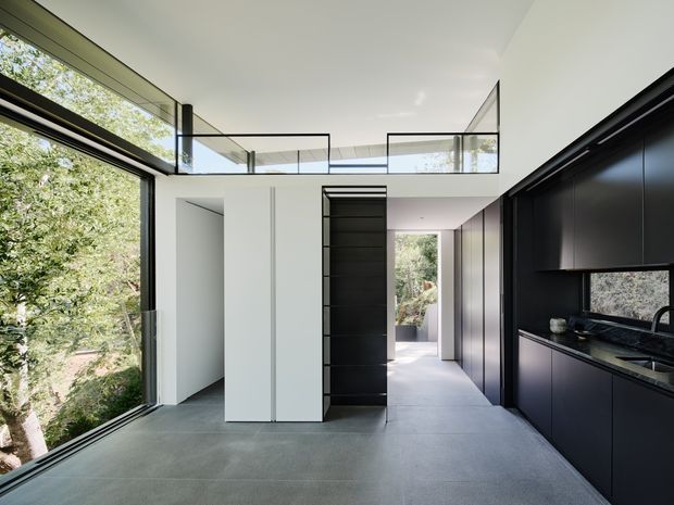 A contemporary open-plan interior with a dark minimalist kitchen along the right wall, integrated black cabinetry, and a large black-framed window overlooking greenery. The space has a pale smooth ceiling and walls, grey tiled flooring, a bright circulation area, and a glass balustrade around an upper-level landing or void. Full-height glazing brings in abundant natural light.