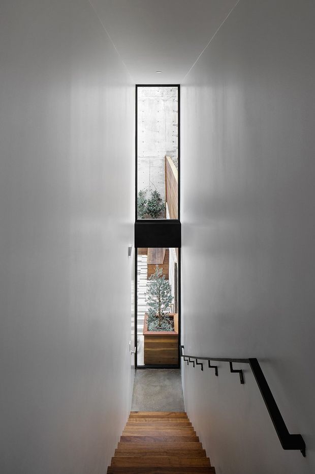 A narrow, minimalist stairway descends between smooth white walls toward tall black-framed glazing. Warm timber stair treads contrast with the cool grey floor and black handrail, while a small planted courtyard and timber-clad exterior elements are visible beyond the window. The composition emphasizes the stair's linear perspective and vertical light well.